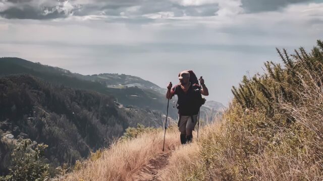 Man with backpack and trekking poles hiking a grassy ridge above forested mountains with sea in the distance under a cloudy sky