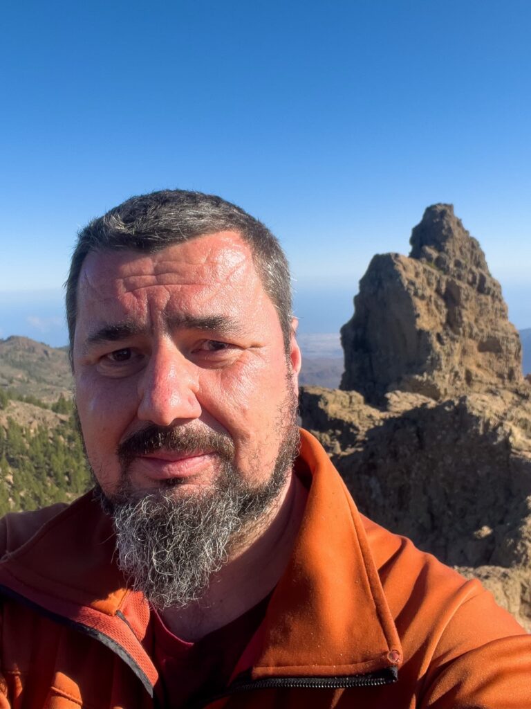 Man with salt-and-pepper beard wearing an orange jacket taking a selfie against a rocky peak and clear blue sky